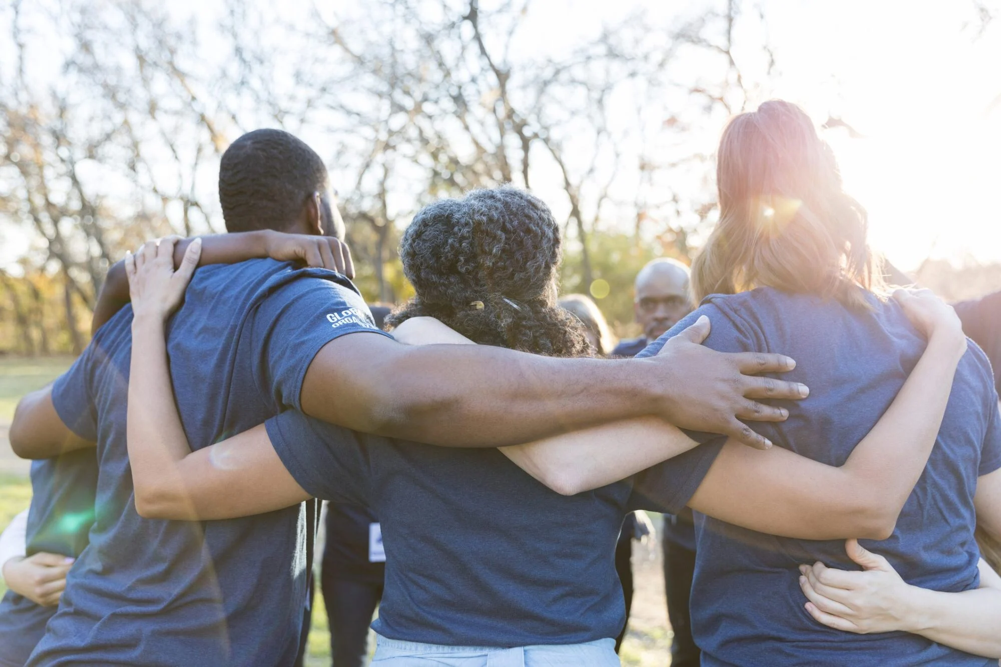 A group of people in blue shirts stand outdoors, arms around each other in a circle, facing away.